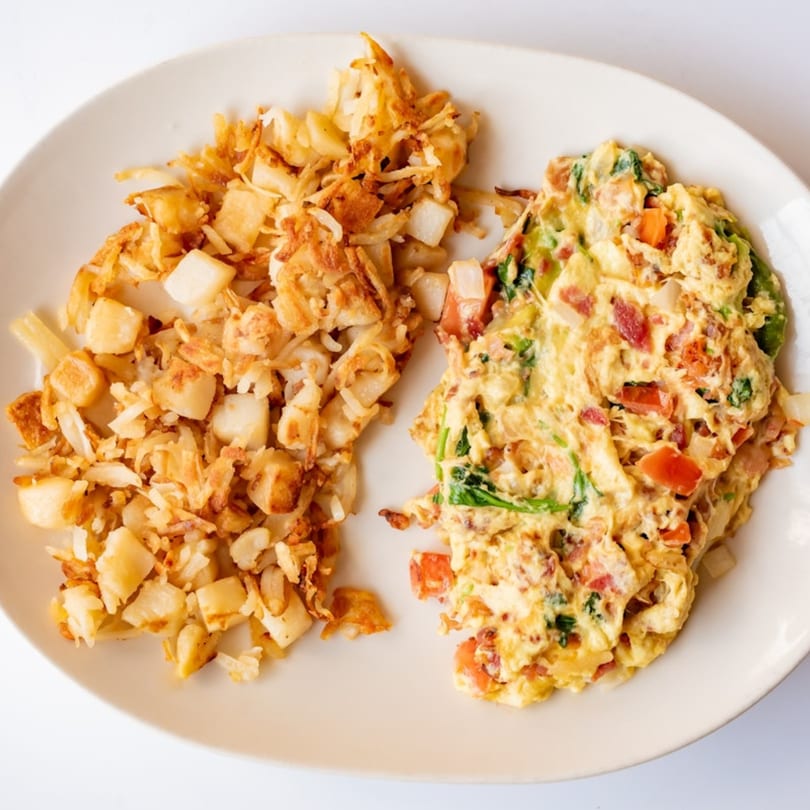 A plate of scrambled eggs with vegetables and bacon, alongside crispy hash browns.