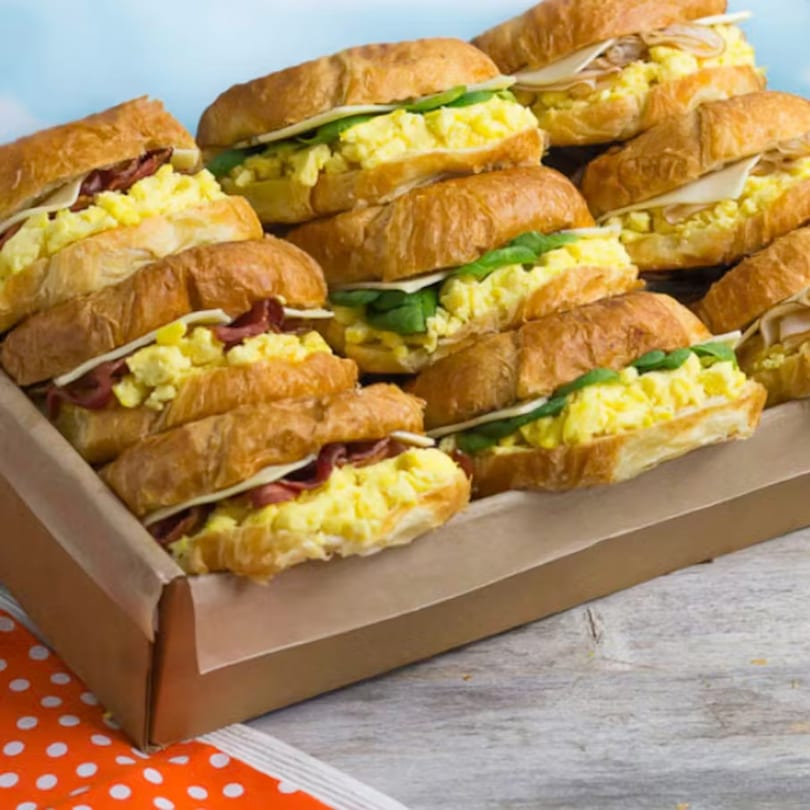 A catering tray filled with numerous croissant sandwiches, some with bacon and others with spinach.