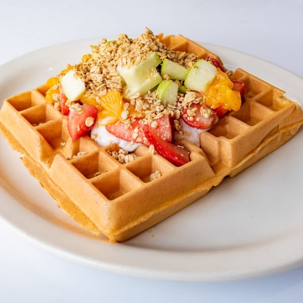 Golden waffle topped with yogurt, fresh strawberries, oranges, green apples, and granola on a white plate.