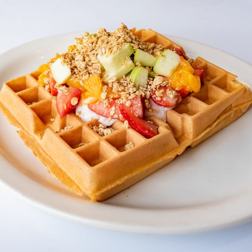 Golden waffle topped with yogurt, fresh strawberries, oranges, green apples, and granola on a white plate.