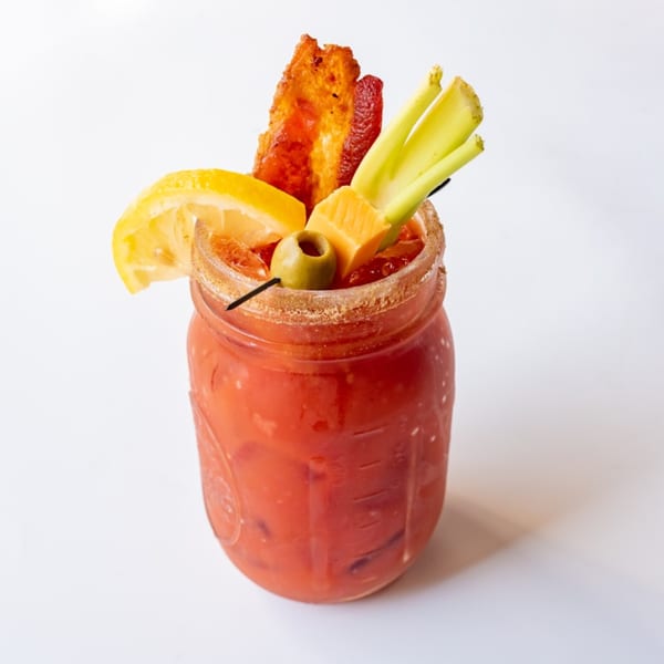 A KupAJoe Cafe Bloody Mary in a mason jar, garnished with bacon, celery, lemon, olive, and cheese.