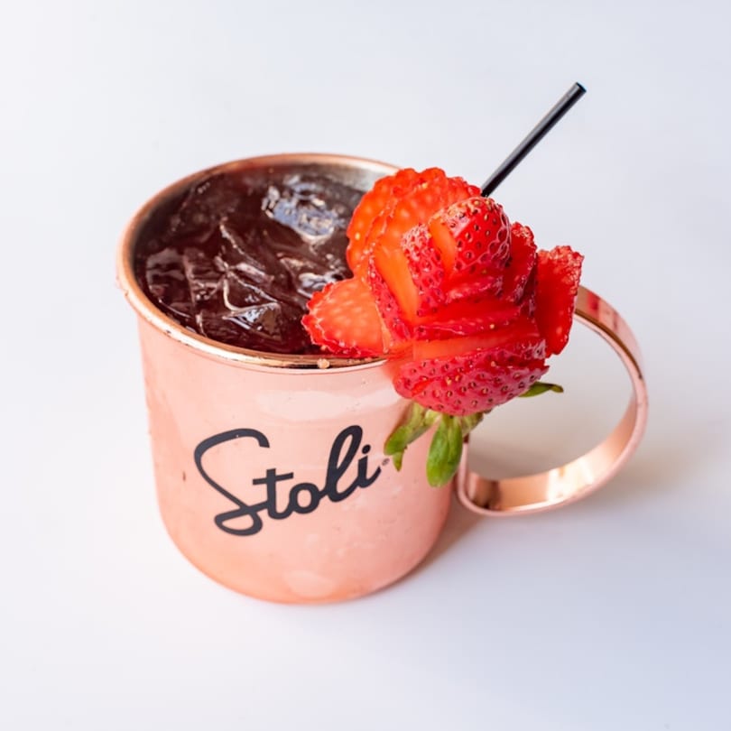 A copper Stoli mug filled with a dark drink and ice, garnished with a strawberry rose.