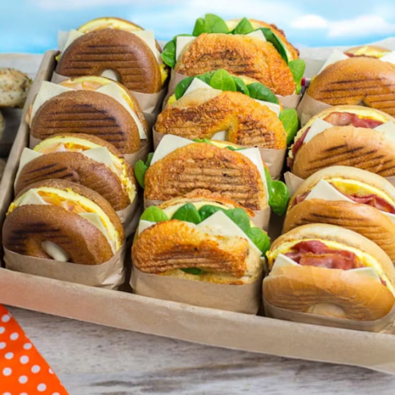 A catering tray filled with various breakfast bagel sandwiches, some with bacon, egg, and cheese.